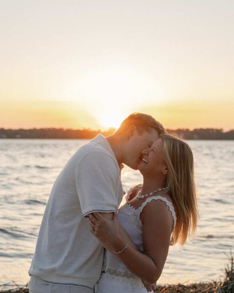 boyfriend whispering something in girlfriends early to make her laugh at the beach on with the ocean and sunset behind them 